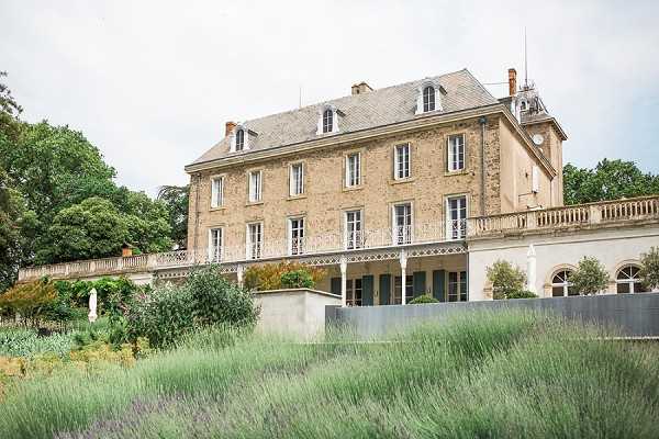 Exterior wide shot of a French chateau photographed from the grounds, showing a three-storey limestone manor house with a slate mansard roof, dormer windows, and white wrought-iron balustrades along a covered terrace on the ground floor. The foreground features rows of lavender plants in full bloom alongside low ornamental plantings, with a modern rectangular stone or concrete planter element visible at mid-ground. No people are present in the image. Potential venue feature image.