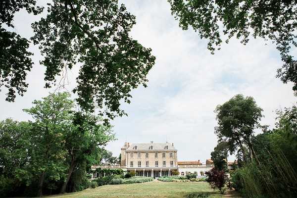 Wide exterior shot of a French chateau viewed from across its grounds, framed by large mature trees in the foreground. The building is a classic two-storey stone manor with symmetrical windows, a mansard roof with dormer windows, and a covered terrace or orangerie wing extending along its facade. No people are visible in the image. The composition is a wide landscape shot taken from a distance, with the chateau positioned centrally in the middle ground. Potential venue feature image.