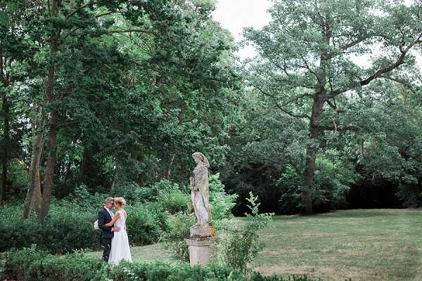 A wide-shot couple portrait taken outdoors in a formal garden setting, with the bride and groom standing close together and facing each other in what appears to be a quiet, intimate moment. The bride wears a white gown and the groom is dressed in a dark navy suit. A weathered stone classical statue on a pedestal stands prominently to the right of the couple, adding a formal garden character to the composition. The surrounding landscape features mature trees with dense green foliage framing the scene.