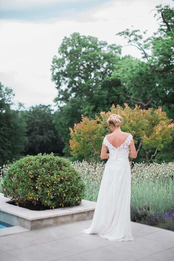 A bridal portrait taken outdoors, showing the bride from behind as she stands on a stone-paved terrace beside a pool. She is wearing a white gown with a low V-back adorned with delicate floral lace appliqué details and a flowing chiffon skirt with a short train. Her blonde hair is styled in an updo. The terrace features a clipped topiary shrub in a raised stone planter, and a row of lavender in bloom borders the garden beyond. The composition is a full-length portrait shot from behind, with the bride facing the garden.