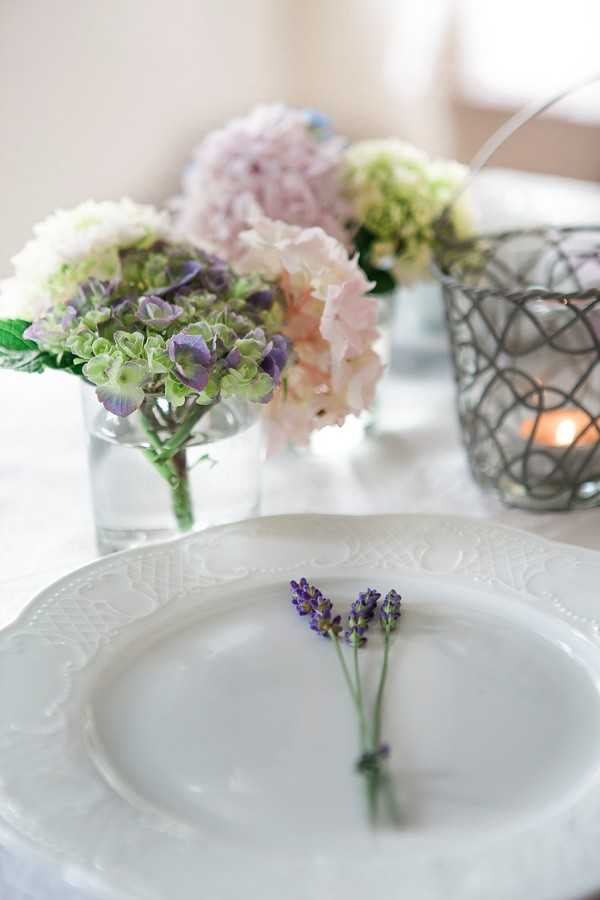 A close-up detail shot of a wedding table setting featuring a white embossed charger plate with two small sprigs of purple lavender tied together placed on top. In the background, multiple low glass vases hold clusters of hydrangeas in blush pink, soft purple, green, and white tones, arranged as centerpieces along a white linen table. A wire lattice votive candle holder with a lit candle sits to the right of the floral arrangements. The overall decor palette is soft and romantic, combining lavender, blush, and white in a French country or garden-party style.