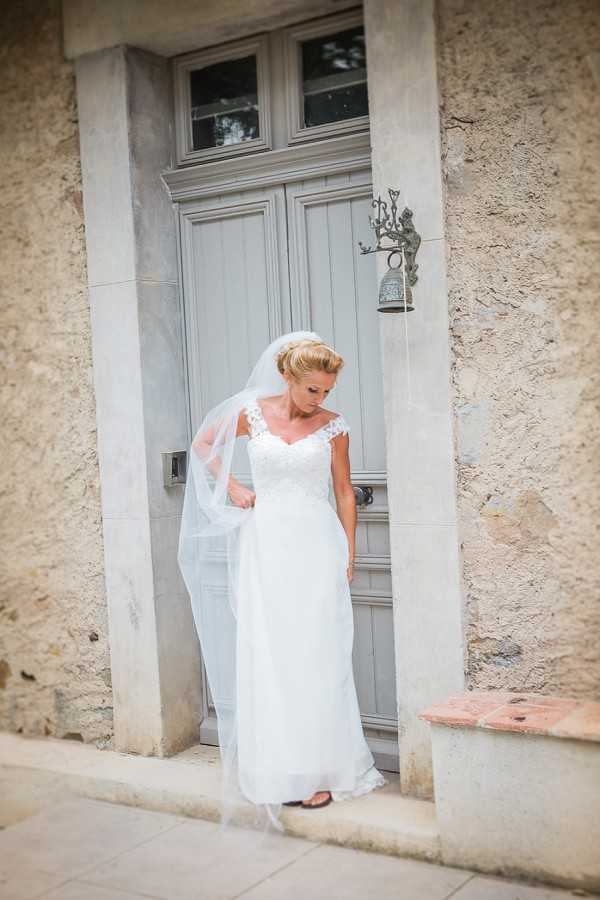 A bridal portrait taken outdoors at what appears to be a French property, with the bride standing in the doorway of a building featuring a pale grey-painted wooden door with glass transom panels and a decorative cast-iron bell fixture. The bride wears a white cap-sleeve gown with a lace bodice and flowing chiffon skirt, paired with a long cathedral-length veil and an updo hairstyle. She is looking downward and holding the edge of her veil with one hand. The composition is a full-length portrait shot with the doorframe used as a natural framing device.