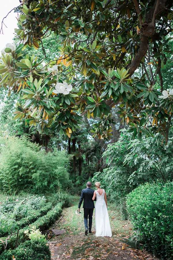 A bride and groom walk away from the camera along a garden path, arms linked, during what appears to be a post-ceremony portrait session in an outdoor garden setting. The groom wears a dark navy suit and the bride wears a white gown with a lace illusion open back, her blonde hair pinned up. A large magnolia tree with white blooms dominates the upper portion of the frame, with dense, manicured greenery lining both sides of the path. The composition is a wide shot taken from behind the couple, emphasizing the lush garden surroundings and the intricate back detailing of the bridal gown.