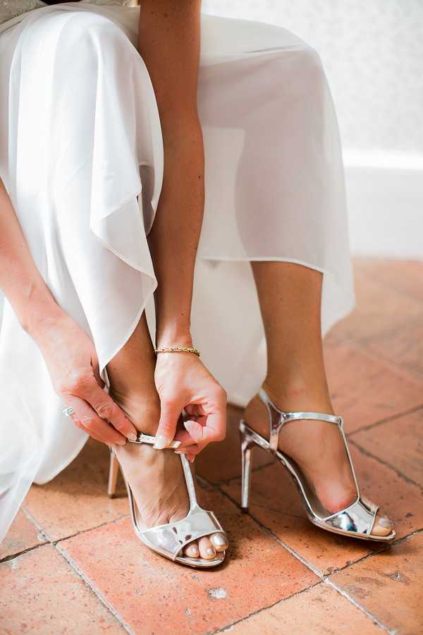 Close-up detail shot of a bride getting ready, fastening the ankle strap on a pair of silver metallic stiletto heeled sandals with a t-bar design. The bride is wearing a white dress with flowing, layered chiffon fabric and has a delicate gold chain bracelet on her wrist. She is seated on a terracotta tiled floor, and a engagement or wedding ring is visible on her finger. The composition is tightly framed on the feet and hands, emphasizing the shoe styling choice against the warm-toned floor tiles.