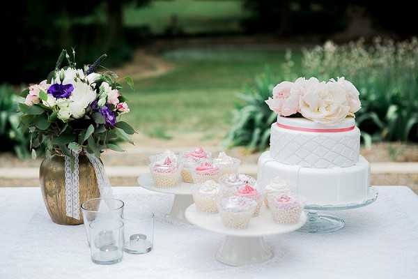 A dessert table styled for a wedding reception, photographed outdoors with a garden backdrop. The table features a two-tier white fondant cake with a diamond quilted pattern, a coral-pink ribbon between the tiers, and large blush pink peonies on top, displayed on a glass cake stand alongside a white pedestal stand holding approximately eight cupcakes decorated with white and pink frosting. To the left sits a bronze/gold vase tied with white lace ribbon, holding a mixed bouquet of white ranunculus, purple anemones, pink spray roses, and eucalyptus foliage, with two small clear glass votive candle holders nearby. The overall decor palette is white, blush pink, coral, and purple with gold accents, suggesting a classic-romantic styling. Wide, slightly shallow depth-of-field shot taken at table level.