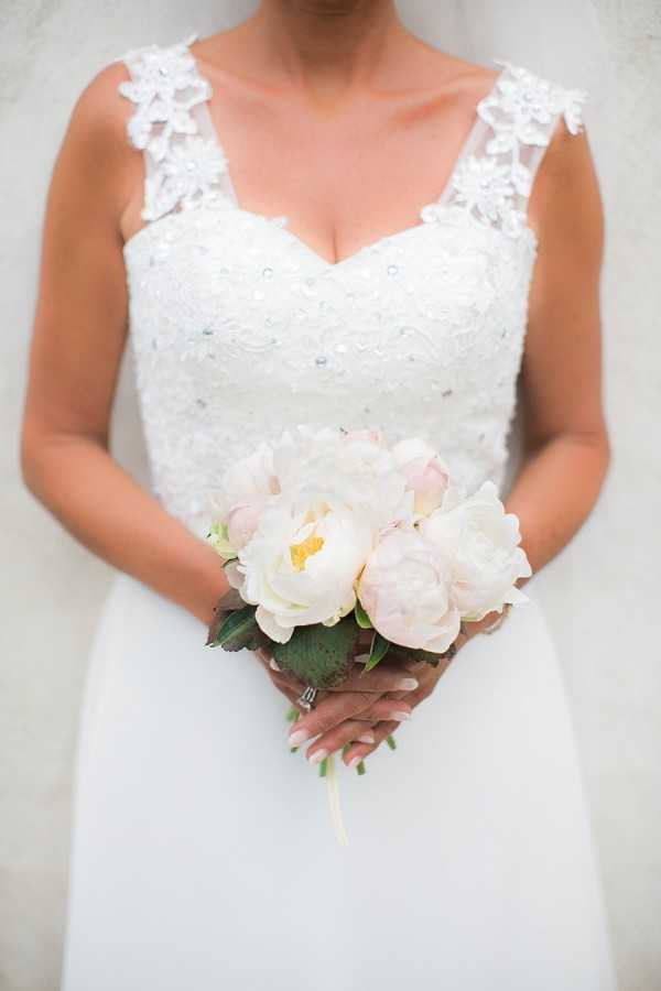 Close-up portrait of a bride from the neck down, holding a small, tightly gathered bouquet of cream and blush peonies with green foliage. The bride wears a white gown with a sweetheart neckline, beaded lace bodice with scattered crystal embellishments, and wide lace appliqué straps featuring floral detailing. A white veil is partially visible behind her shoulders. Her nails are painted with a French manicure and she wears a ring on her right hand. The bouquet is tied with a pale ribbon at the stems.