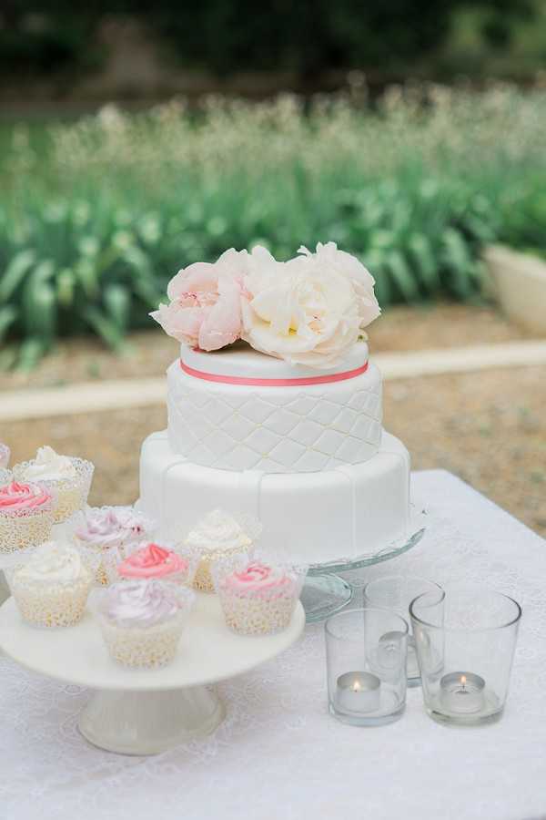 Close-up detail shot of an outdoor dessert table styled in a soft, feminine palette. The centerpiece is a two-tier white fondant wedding cake with a quilted diamond pattern on the upper tier, a coral-pink ribbon accent between tiers, and blush and cream peonies as a floral topper, displayed on a glass cake stand. To the left, a white pedestal stand holds an assortment of cupcakes with pink, lavender, and ivory frosting in lace-edged wrappers. Two small glass votive candle holders with lit tea lights are placed to the right of the cake. The table is covered in a white lace-trimmed cloth, and the setting appears to be an outdoor garden or terrace.