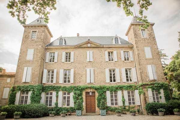 Wide exterior shot of a French chateau with cream-colored stone façade, pale blue-grey shutters, and twin tower wings flanking the central three-story main building. The entrance is framed by climbing green ivy and features a large wooden double door, with potted topiaries and small teal planters arranged along the gravel forecourt. The architecture includes a clock medallion above the central doorway and slate-tiled mansard roofs on the towers. Potential venue feature image.