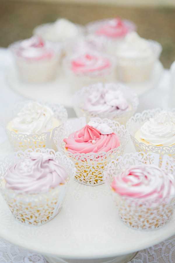 Close-up detail shot of decorated wedding cupcakes displayed on a white cake stand with a lace doily. The cupcakes are wrapped in white and gold laser-cut lace cupcake wrappers and are frosted in a palette of bright pink, soft lavender, and ivory white swirled buttercream, some topped with small sugar flower decorations and pearl sprinkles. A second cake stand with additional cupcakes is visible in the soft-focus background. The overall dessert table styling follows a romantic, feminine theme with a pink, lavender, and white color palette.