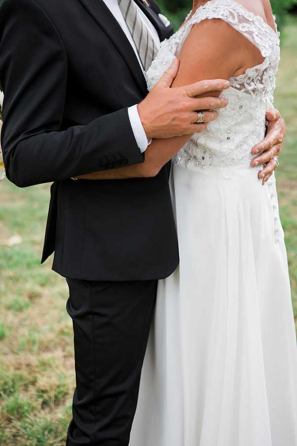 A close-up portrait of a couple posing outdoors on grass, cropped to show their torsos only. The groom wears a dark navy or black suit with a grey striped tie and white dress shirt, with a gold wedding band visible on his left hand as he rests it on the bride's back. The bride wears a white gown with a lace and beaded cap-sleeve bodice featuring delicate embroidery and small buttons down the back, with a flowing chiffon skirt. The composition is a tight mid-section detail shot that highlights the groom's hand placement and the intricate lace detailing on the dress back.