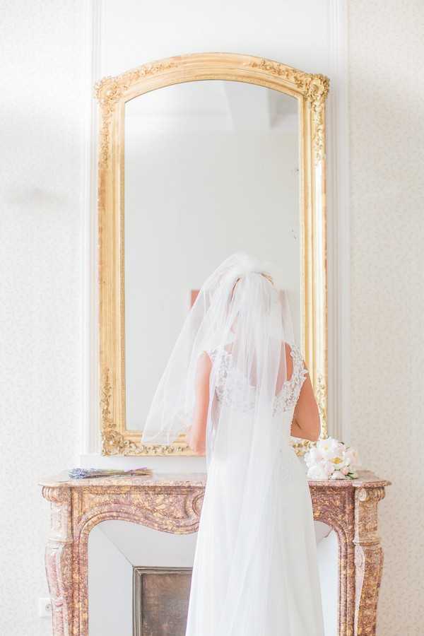 A bride stands with her back to the camera during a getting-ready moment, facing a large ornate gold-framed mirror mounted above a pink and gold marble fireplace mantel. She is wearing a white gown with a lace-detailed back and a two-tier white cathedral-length veil that cascades down her back. A small bouquet of white blooms, likely peonies or garden roses, rests on the right side of the mantel, and a small bunch of dried lavender sits on the left. The interior setting features soft white walls with subtle wallpaper texture, consistent with a classic French chateau style. The shot is a medium portrait taken from behind, with bright, airy natural lighting.
