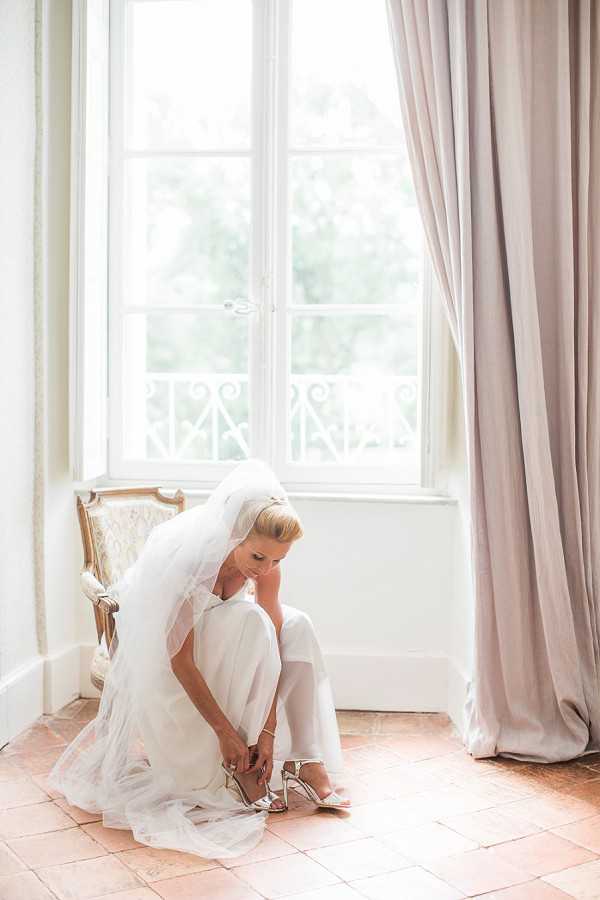 A bride is photographed getting ready, fastening a strappy silver heel while seated near a large French window with white-painted frames and wrought-iron balcony details. She is wearing a white bridal jumpsuit and a long, flowing white tulle veil, with her blonde hair styled up. The room features terracotta tile floors, a gold-framed French-style upholstered chair, and floor-length taupe curtains, suggesting a chateau or manor house interior. The image is a full-length portrait shot, lit by soft natural light flooding through the window.