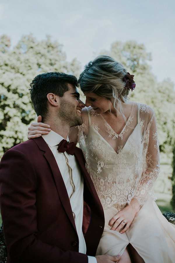 A couple portrait taken outdoors, with the bride seated and leaning forehead-to-forehead with the groom, both laughing. The groom wears a deep burgundy suit jacket with a matching burgundy bow tie and an ivory dress shirt. The bride wears a long-sleeve lace overlay wedding dress with a V-neckline in ivory and blush tones, and has a dark burgundy/plum floral hair accessory tucked into a loose updo. The color palette of burgundy and blush is consistent across both outfits, suggesting a coordinated boho-romantic styling theme. The shot is a close-up portrait with a shallow depth of field, taken in a natural outdoor setting.