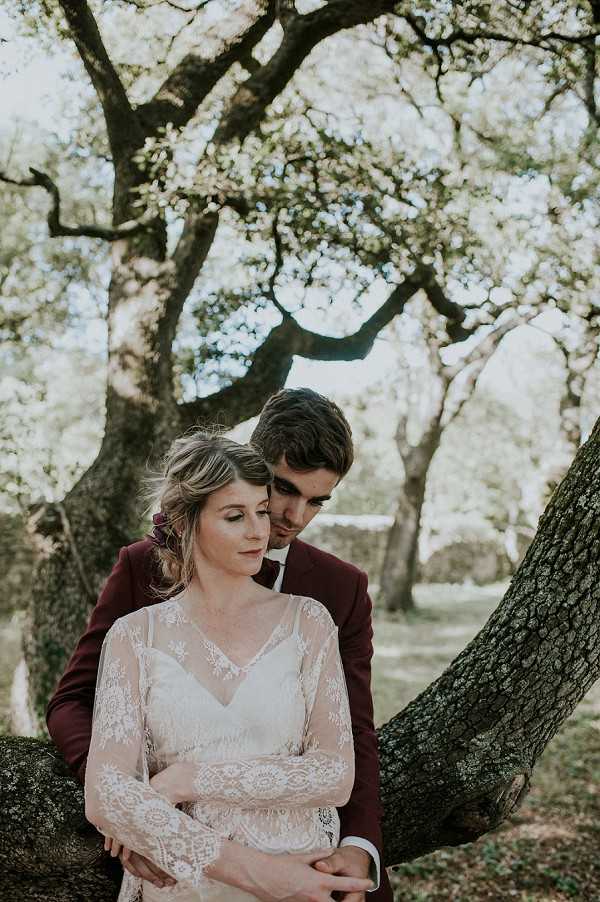 A couple portrait shot outdoors among large trees, with the groom standing behind the bride and embracing her from behind. The bride wears a long-sleeve ivory lace dress with intricate floral lace detailing and a V-neckline, with her hair loosely pinned up and decorated with a small dark burgundy floral accent. The groom wears a deep burgundy suit jacket with a dark bow tie. The setting is a wooded outdoor area, and the styling leans toward a boho-romantic aesthetic with the pairing of lace and rich jewel tones. The composition is a mid-range portrait with soft, diffused natural backlighting filtering through the tree canopy.