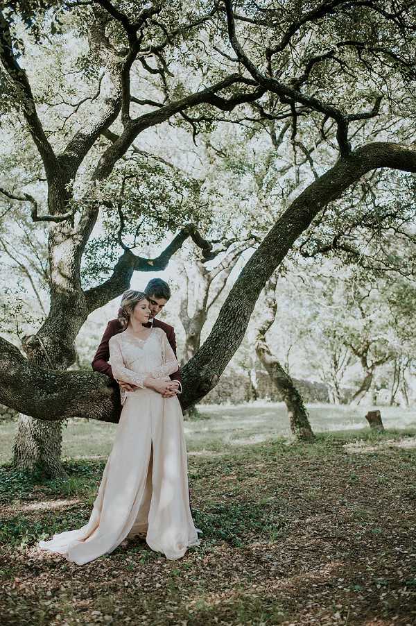 A couple portrait taken outdoors beneath a large, spreading oak tree with gnarled branches. The groom, wearing a deep burgundy suit, stands leaning against the tree trunk with his arms wrapped around the bride from behind, his head bowed toward hers. The bride wears a long-sleeved lace-bodice gown in ivory with a flowing satin skirt featuring a front slit and a trailing hem. Her hair is styled in a loose updo. The composition is a medium-wide portrait shot with the tree's sweeping branches framing the couple from above, set within a wooded area with dappled natural light filtering through the canopy.