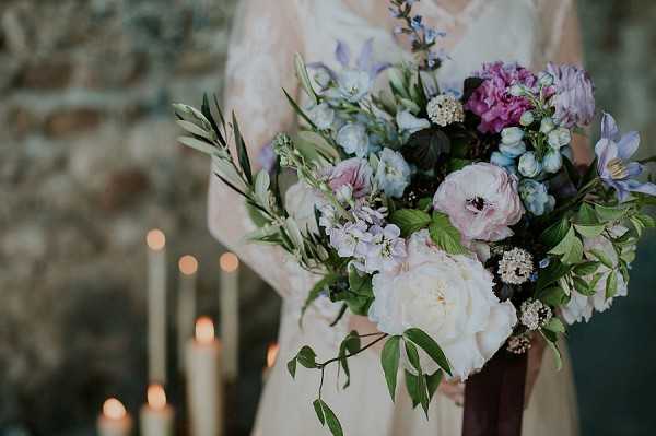 Close-up detail shot of a bridal bouquet being held by a bride wearing a lace long-sleeved ivory dress. The loose, garden-style bouquet features white peonies, blush ranunculus, mauve scabiosa, blue delphinium, purple hydrangea, pale blue nigella, and trailing olive and eucalyptus greenery, tied with a deep burgundy ribbon. In the soft-focus background, a row of tall lit taper candles in gold holders is visible against a stone wall, suggesting a rustic or historic indoor venue setting.