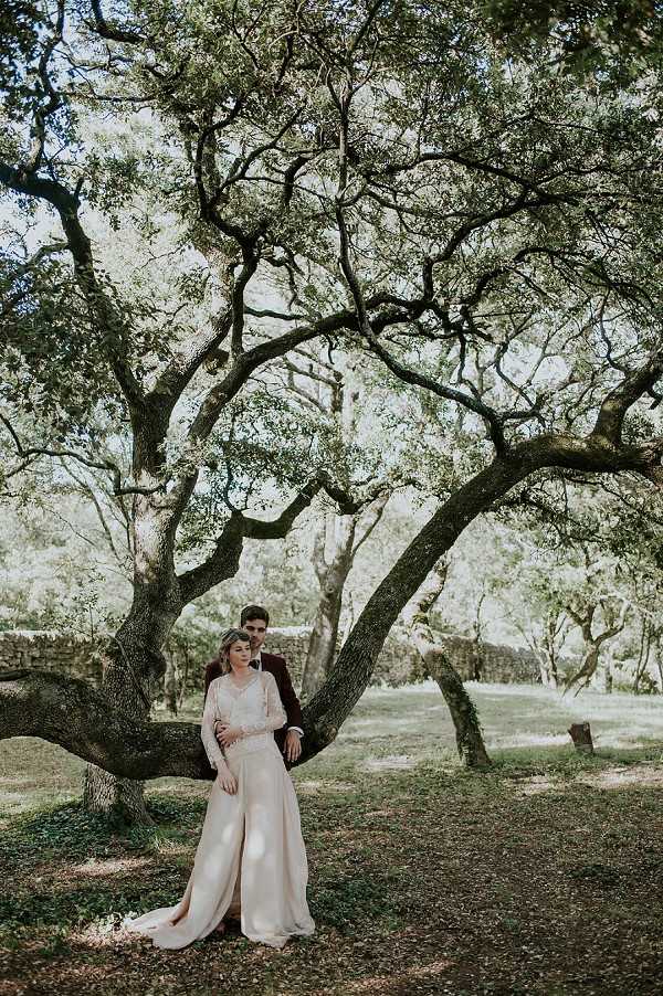 A couple portrait taken outdoors beneath a large, sprawling tree with wide-reaching branches in a wooded parkland setting. The bride wears a long-sleeved ivory gown with a lace bodice and a flowing skirt with a train, while the groom is dressed in a deep burgundy suit. The groom stands slightly behind the bride, leaning against the tree trunk with his arms around her as she leans back into him. The composition is a full-length portrait with the tree canopy filling most of the frame, shot in natural dappled light. The styling has a romantic, bohemian feel, and a low stone wall is faintly visible in the background.