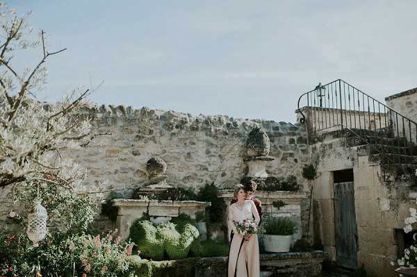 A bride stands alone in an outdoor courtyard garden against an aged stone wall with decorative stone urns and a weathered teal-painted door. She is wearing a long-sleeved blush pink gown and holding a loose, garden-style bouquet featuring mauve, dusty pink, and white blooms with greenery. The courtyard features terracotta pots, trimmed hedges, and a wrought-iron staircase railing, suggesting a rustic Provençal mas or historic stone property. The shot is a wide portrait composition with the bride positioned slightly off-center amid the lush planted surroundings.