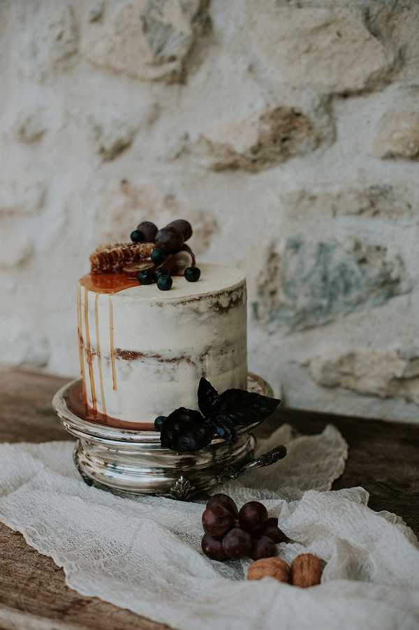 Close-up detail shot of a single-tier semi-naked wedding cake displayed on a silver cake stand, set against a rustic exposed stone wall. The cake features a caramel drip finish, topped with a honeycomb piece, fresh blueberries, and dark chocolate or licorice sticks, with dark purple-black decorative leaves and blueberries at the base. The cake stand rests on a wooden surface draped with ivory cheesecloth fabric, with a small cluster of dark red grapes and three walnuts arranged in the foreground as styling props. The overall aesthetic is rustic and organic, with a warm, earthy color palette of ivory, caramel, deep navy-purple, and silver.