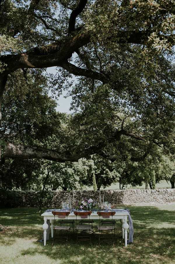 An outdoor reception tablescape set beneath a large spreading tree in a garden or domaine grounds, with no people present. The white-painted farmhouse-style dining table is dressed with a pale grey linen runner and set for approximately six guests using dark metal bistro-style chairs. Table decor includes copper or terracotta-toned charger plates, glassware, tall thin taper candles, and a central floral arrangement featuring purple and mauve blooms. The styling palette combines muted blues, copper, and soft purples for a rustic-romantic aesthetic. A dry-stone wall runs across the background. Wide shot taken from ground level, capturing the full table within its outdoor setting.