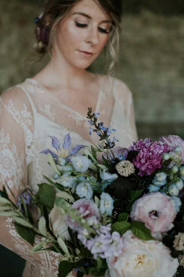 A close-up bridal portrait of a bride looking down at her bouquet, with the flowers in sharp focus in the foreground. The bride wears an ivory lace gown with sheer long sleeves and detailed floral lace patterning, and her hair is loosely pinned up with small purple flowers tucked in. The large, loosely arranged bouquet features blush and mauve peonies, cream garden roses, pale blue clematis, blue delphinium, white scabiosa, blackberries, and lush green foliage, creating a wild, romantic boho aesthetic. The overall color palette leans cool — blues, purples, and soft pinks — against the warm ivory of her dress.