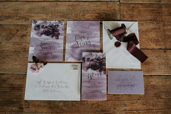 A flat lay detail shot of a wedding stationery suite arranged on a rustic wooden surface. The collection includes two invitation cards, a menu card, an RSVP card, and two envelopes — one addressed in white calligraphy script and one sealed with a deep burgundy wax seal. The stationery features a watercolor wash background in muted mauve and dusty purple tones, accented with dark botanical illustrations of roses and foliage in deep plum and grey-purple hues. A spool of dark burgundy silk ribbon is placed alongside the sealed envelope, and a small dried or silk dusty rose bloom is tucked near one of the invitations. The overall palette is moody and romantic, combining deep wine, mauve, and soft grey tones in a modern calligraphy style.