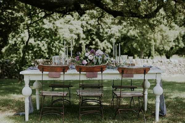 A styled outdoor reception tablescape set on a lawn beneath a large tree, with no people present. The table is a distressed white farmhouse-style piece flanked by vintage wrought-iron bistro chairs with slatted wooden seats. The centerpiece features a wooden bowl filled with purple, lavender, and blush florals including what appear to be ranunculus and wildflowers, accompanied by deep purple blooms and trailing greenery. Tall thin taper candles in soft white are spaced along the table, and crystal glassware including champagne flutes is arranged at each place setting. Two terracotta-style wooden bowls flank the central arrangement, and a dusty blue-grey linen runner drapes loosely along the table. The overall styling palette is rustic-romantic with a muted lavender, blush, and natural wood tone scheme. Wide establishing shot of the full table setup.