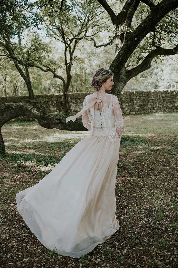 A bridal portrait taken outdoors in a wooded garden setting, showing the bride from behind and in three-quarter profile. She wears a champagne-toned ball gown with a flowing skirt and a sheer lace cape or bolero featuring long bell sleeves and a satin bow tied at the back. Her hair is styled in an updo with small deep purple floral accents. The skirt billows outward, suggesting movement. The overall styling has a boho-romantic aesthetic. Medium full-length shot.
