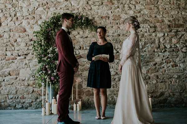 A wedding ceremony is taking place indoors against an exposed stone wall, with the couple facing each other and an officiant standing between them holding a booklet. The groom wears a deep burgundy suit with a bow tie, and the bride wears a long ivory dress with lace long sleeves and her hair pinned up with a floral accent. Behind them is a large circular greenery arch decorated with pink and mauve flowers, flanked by clusters of white pillar candles in varying heights arranged on the concrete floor. The officiant wears a navy blue knee-length dress. The setting has a rustic feel with the raw stone wall backdrop and polished concrete floor. The image is a medium wide shot capturing all three figures full-length.
