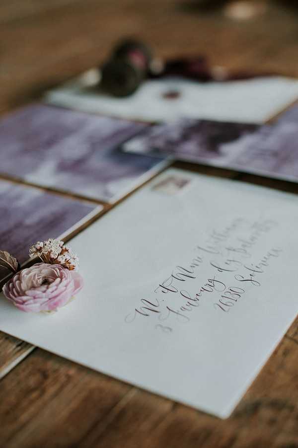 Close-up detail shot of a wedding stationery flat lay arranged on a rustic wooden surface. The suite includes a white envelope addressed in handwritten calligraphy script in a dark ink, accompanied by cards or inserts featuring moody mauve and purple watercolor wash designs. A single dusty pink ranunculus bloom with small dried white filler flowers is placed alongside the envelope as a styling accent. A wax seal is partially visible in the background. The overall palette is muted — soft whites, dusty pinks, and deep plum purples — suggesting a romantic, moody stationery aesthetic.