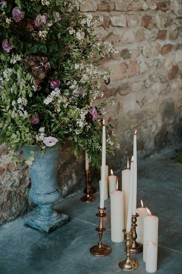 A close-up detail shot of a wedding floral and candle arrangement placed on a concrete floor against a rustic exposed brick wall. A weathered blue-grey stone urn holds an overflowing arrangement of mauve ranunculus, soft lavender anemones, small white blossoming branches, and lush green foliage. Alongside the urn, a cluster of lit ivory taper candles in antique brass candlesticks and several pillar candles are grouped together on the floor, casting a warm glow. The overall decor palette combines dusty purples, aged brass, and muted grey-blues, with a romantic, rustic-classical styling theme.