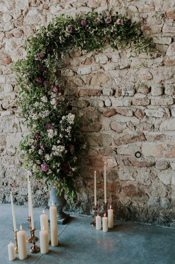 A ceremony or styled decor detail shot featuring a large circular floral arch displayed against an exposed stone wall indoors. The arch is densely arranged with lush greenery, small white blooms, and deep purple and mauve roses, creating a wild, organic aesthetic. At the base of the arch, a collection of cream pillar candles and tapered candles in brass and copper candlesticks are arranged on a polished concrete floor, with several candles lit. The overall decor palette combines deep plum, cream, and green with warm metallic accents, suggesting a romantic rustic styling theme.