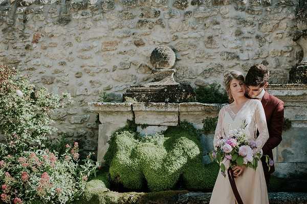 A couple poses together outdoors in front of an aged stone wall with a decorative stone fountain or pedestal covered in moss. The groom stands behind the bride, leaning in close to her cheek, with his arms around her; he wears a deep burgundy suit. The bride wears a white long-sleeve dress with a V-neckline and holds a loose bouquet of soft pink peonies and lilac blooms with green foliage. The overall styling is romantic and classic with a slightly rustic feel, complemented by the weathered stone backdrop. The shot is a medium portrait framing both figures from approximately the knees up.