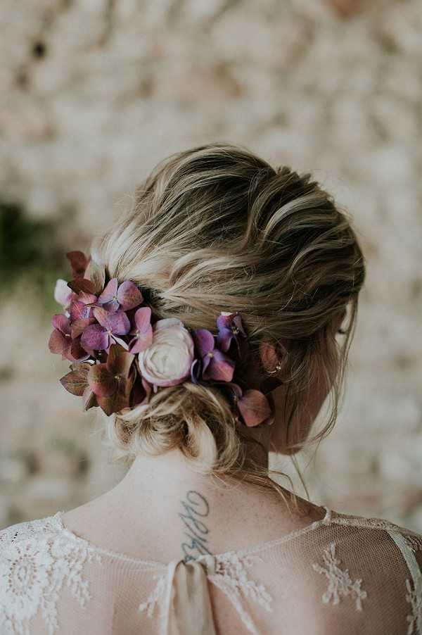 Close-up portrait shot from behind of a bride with a loosely pinned updo styled with fresh flowers, including dusty mauve and purple hydrangea petals and a cream ranunculus bloom. The bride is wearing a sheer lace-detailed dress with a blush or ivory tone, and a small tattoo is visible on her upper back. A small gold hoop earring is visible on her ear. The background appears to be a floral wall installation with soft, blurred tones. The overall styling aesthetic is boho with a muted, earthy color palette.