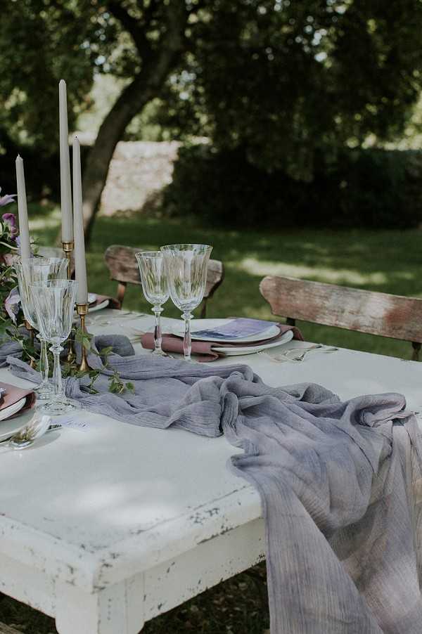 Close-up detail shot of an outdoor wedding reception table setting, styled in a rustic romantic theme. The white distressed-paint wooden table is dressed with a loosely draped dusty blue-grey cheesecloth gauze runner, layered with trailing greenery and purple sweet pea florals. Place settings include stacked terracotta and blush ceramic plates topped with mauve linen napkins, crystal cut-glass wine goblets, and small calligraphy place cards, with brass candlestick holders holding tall taupe taper candles. The seating consists of weathered, peeling wooden garden benches, reinforcing the rustic outdoor aesthetic.