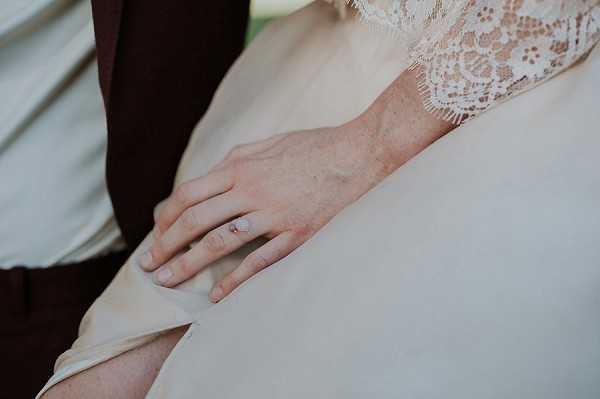 Close-up detail shot of a bride's hand resting on the skirt of her ivory wedding dress, showing a delicate solitaire engagement ring with a small round stone on a thin band. The bride's lace bodice with scalloped edges is visible in the upper portion of the frame, and the groom's dark burgundy suit sleeve is partially visible to the left. The soft, smooth fabric of the bridal skirt contrasts with the textured lace of the bodice, suggesting a classic bridal style.