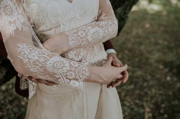 Close-up detail shot of a couple embracing outdoors, with the groom's arms wrapped around the bride from behind. The bride is wearing a long-sleeve ivory lace wedding dress featuring intricate floral and geometric lace patterns with scalloped cuffs, layered over what appears to be a champagne or blush underlining. The groom is wearing a dark burgundy jacket with a white dress shirt cuff visible. The composition is tightly cropped at torso level, focusing entirely on the textile detail of the lace sleeves and the couple's clasped hands.
