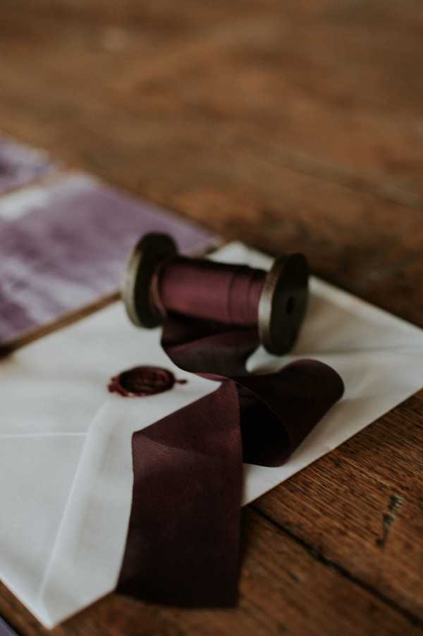 A close-up detail shot of wedding stationery styling laid on a rustic wooden surface. A cream vellum envelope sealed with a small dark red wax seal is the central element, with deep burgundy/plum silk ribbon unspooled across it from a wooden bobbin spool. A second spool of the same ribbon sits nearby, and a blurred mauve or dusty purple silk ribbon is visible in the background. The overall palette is deep burgundy, plum, and ivory, suggesting a moody, romantic styling direction. The shot is taken at a shallow depth of field, with soft, low natural light creating warm, dark tones throughout.