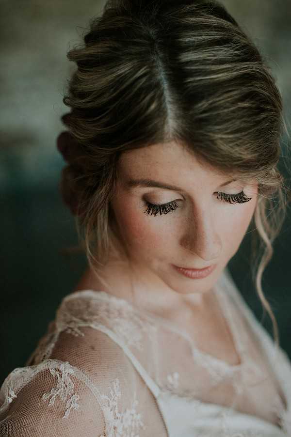 Close-up portrait of a bride looking downward, shot with a shallow depth of field that keeps her face sharp against a blurred dark background. She wears a white lace dress with sheer floral-patterned sleeves and delicate lace detailing at the shoulder. Her hair is styled in a voluminous twisted updo with soft face-framing curls, and her makeup features prominent false lashes, soft blush tones, and a nude lip. The overall styling aesthetic is romantic and vintage-inspired.