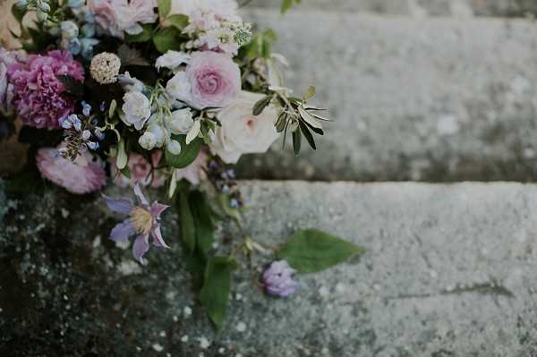 Close-up detail shot of a bridal bouquet resting on aged stone steps. The bouquet features a loose, garden-style arrangement of blush and dusty pink roses, mauve ranunculus, pale blue delphinium, soft white blooms, purple clematis, and olive branch foliage with small leaves trailing downward. A single small purple flower bud has fallen onto the stone surface below. The overall palette is soft and romantic, combining blush, mauve, lilac, pale blue, and ivory tones with deep green foliage accents.