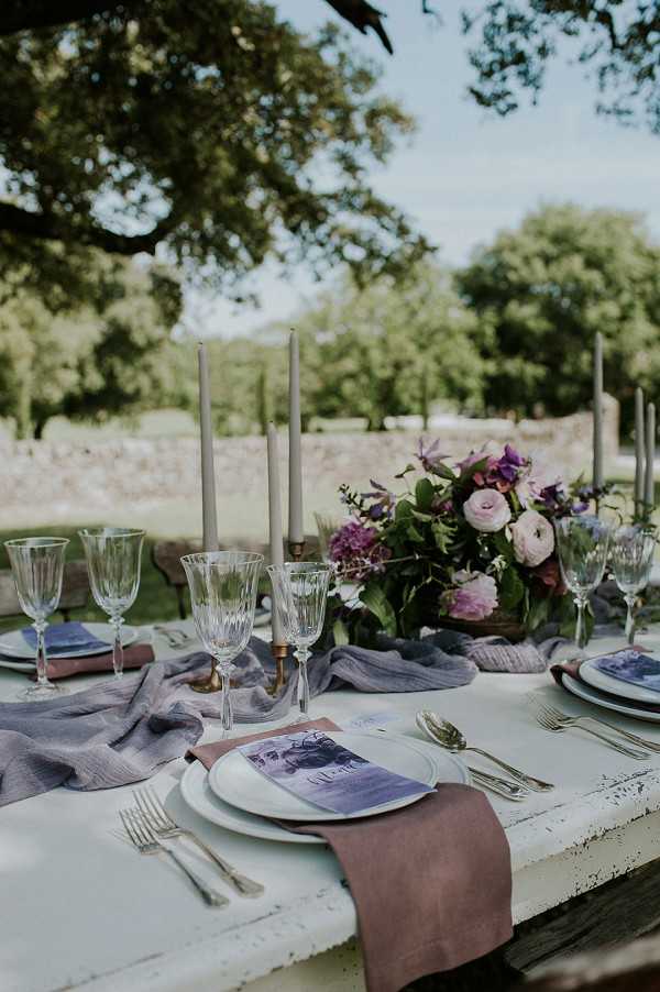 A close-up detail shot of an outdoor wedding reception tablescape set on a whitewashed rustic wooden table. The place settings feature white ceramic plates layered with dusty mauve linen napkins and illustrated menu cards in shades of blue-purple, placed atop a gauzy lavender table runner. Cut crystal wine glasses and gold candlestick holders with tall taupe taper candles are arranged along the center of the table. A floral centerpiece in the background contains blush ranunculus, deep purple flowers, lilac blooms, and lush greenery. The overall decor palette combines dusty mauve, lavender, and deep violet with gold accents, suggesting a romantic, slightly bohemian styling. The table is set outdoors in what appears to be a garden or chateau grounds setting.