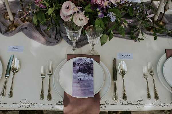 A close-up overhead shot of a styled wedding reception tablescape on a white distressed wooden table. Each place setting features a white charger plate topped with a mauve linen napkin and a watercolor-style menu card in shades of purple and grey with calligraphy script. Gold flatware is arranged on either side of the plates, accompanied by crystal wine glasses and white place cards with handwritten guest names. A lush floral centerpiece runner runs along the center of the table, incorporating blush pink ranunculus, soft purple blooms, and trailing greenery, accented by a draped grey gauze fabric runner and thin brass candlesticks.