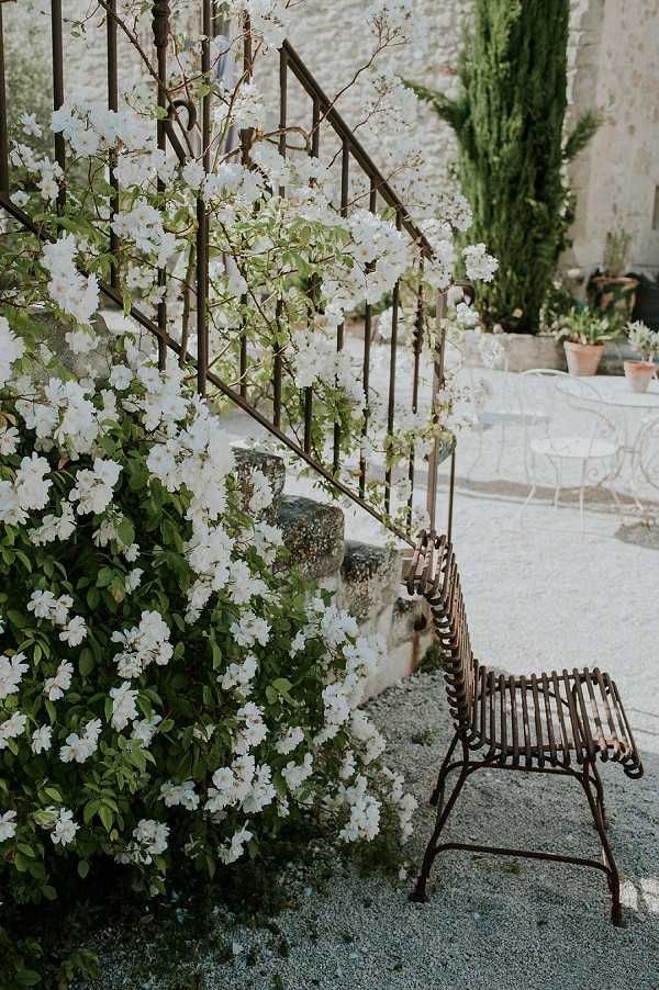 An outdoor venue detail shot featuring a stone staircase with a wrought iron railing covered in a climbing white-flowering shrub in full bloom. A rustic iron slatted chair sits at the base of the stairs on a gravel courtyard. In the background, white wrought iron garden furniture, terracotta planters, and tall cypress trees are visible against a stone building facade. No people are present. The composition is a medium-wide shot with a soft, airy quality. Potential venue feature image.