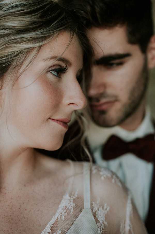 Close-up portrait of a bride and groom posed together, with the bride sharply in focus in the foreground and the groom softly blurred behind her. The bride wears a delicate lace wedding dress with floral lace detailing visible at the neckline and shoulders, and her hair is loosely styled with soft waves. The groom wears a light grey suit jacket with a white shirt and a deep burgundy bow tie. Both are looking downward with calm expressions. The shallow depth of field and warm, soft lighting give the image an intimate, editorial quality.