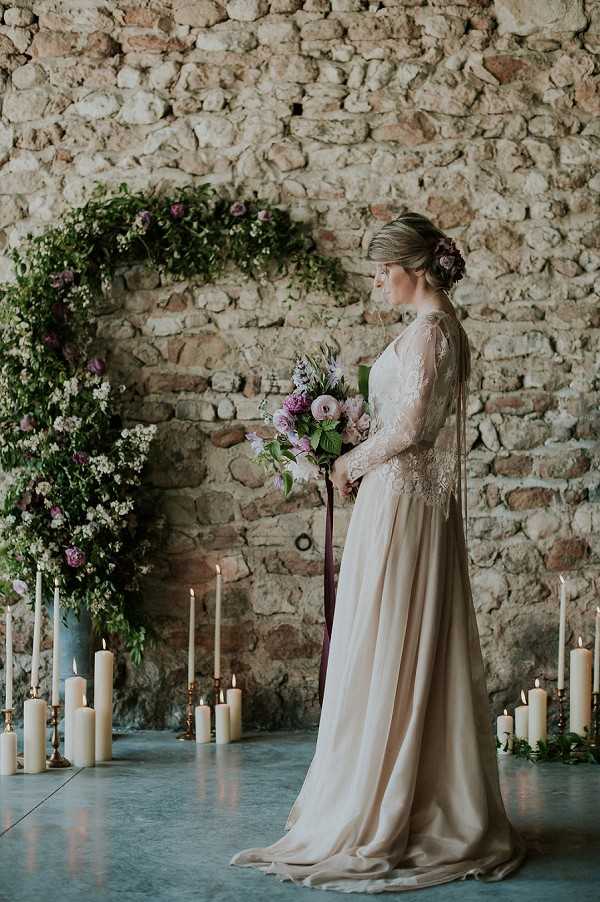 A bridal portrait shot indoors against an exposed stone wall, with the bride standing in profile holding a loose bouquet of blush pink ranunculus, mauve blooms, and greenery tied with a deep burgundy ribbon. She wears a champagne-toned gown with long lace sleeves and a flowing satin skirt, with floral hair accessories pinned into an updo. Behind her, a large circular floral arch constructed from lush greenery, white filler flowers, and purple blooms frames the scene. The floor around her is lined with clusters of ivory pillar candles and taper candles on brass holders at varying heights, creating warm candlelit ambiance. The styling theme is romantic and rustic, combining raw stone architecture with a soft, muted color palette of champagne, mauve, burgundy, and green. Full-length portrait composition.