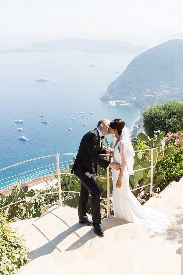 A couple shares a kiss during a portrait session on an outdoor terrace overlooking a deep blue coastal bay with boats and mountains in the background, consistent with the French or Italian Riviera. The groom wears a dark navy suit with a black tie, and the bride wears a fitted white gown with a long flowing veil and a low back. The terrace features a white metal railing and large prickly pear cacti in the foreground. The shot is taken from a slightly elevated angle, capturing the full length of both figures and the dramatic seascape behind them. Potential venue feature image.