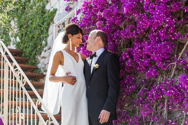 A couple portrait taken outdoors beside a large vibrant bougainvillea in deep magenta-purple in full bloom, climbing a stone wall next to a terracotta-tiled staircase with a white wrought-iron railing. The bride wears a sleeveless, V-neck ivory column gown with a long veil and her dark hair is styled up; the groom wears a classic black tuxedo with a black bow tie and a small white boutonniere. The two are facing each other and smiling closely, suggesting an intimate moment just before or after the ceremony. The shot is a medium portrait with a shallow depth of field that keeps both subjects and the floral backdrop in focus.
