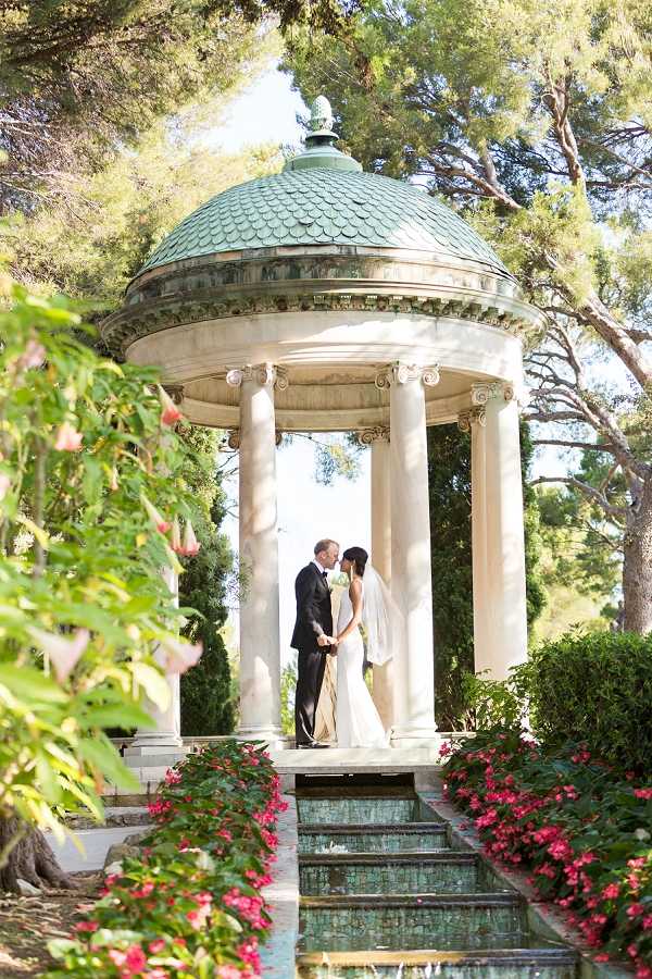 A couple shares a kiss during outdoor wedding portraits inside a circular neoclassical rotunda with white Ionic columns and a green patinated copper dome roof. The bride wears a fitted ivory gown with a long veil, and the groom is dressed in a black tuxedo with a light-colored tie. The rotunda is approached by stone steps flanked by a shallow reflecting pool with a teal-tiled surface and beds of vivid pink flowers on either side. The composition is a medium-wide portrait shot framed through flowering foreground foliage, giving depth to the scene. Potential venue feature image.