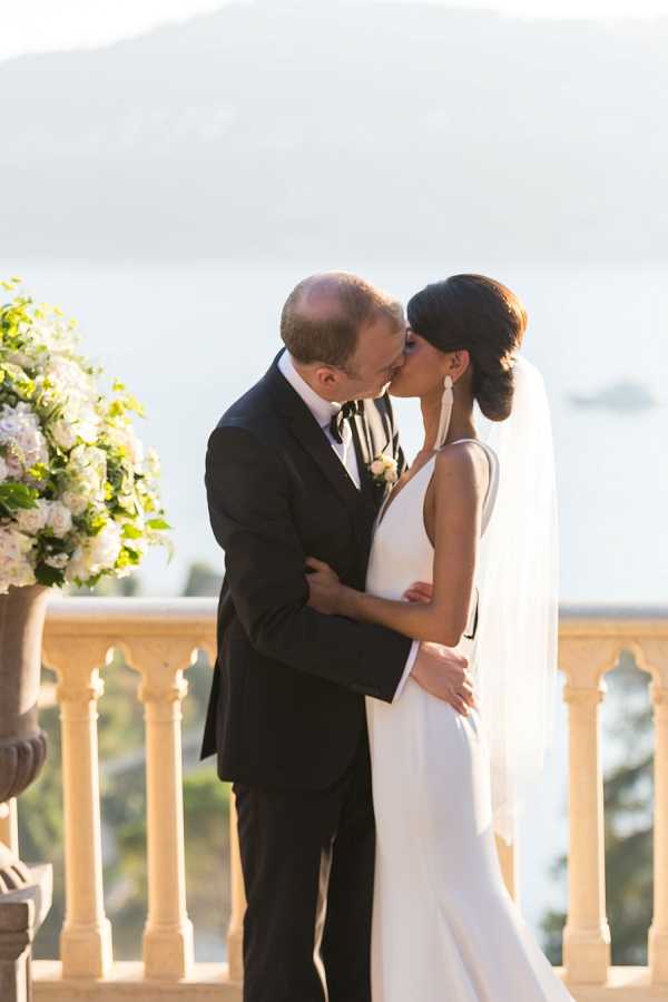 A couple shares their first kiss during an outdoor ceremony on a stone balustrade terrace overlooking a body of water. The groom wears a black tuxedo with a bow tie and a white floral boutonniere, while the bride wears a sleek, minimalist ivory slip-style gown with a long veil and statement white drop earrings, her dark hair styled in an updo. To the left, a large stone urn holds an arrangement of white roses and green foliage. The shot is a medium portrait framing the couple from the waist up, with the classical stone balustrade and water visible in the soft-focus background, suggesting a French Riviera or coastal villa setting with a classic, modern aesthetic.