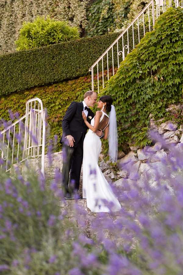 A couple shares a kiss during outdoor portrait photos on stone steps flanked by white iron railings and ivy-covered walls. The bride wears a sleek, form-fitting white gown with an open back and a fingertip-length veil, while the groom is dressed in a black tuxedo with a black bow tie and a small white boutonniere. The foreground is filled with blurred purple lavender blooms, creating a soft frame around the subjects. The composition is a mid-distance portrait shot with shallow depth of field, styled in a modern, clean aesthetic.